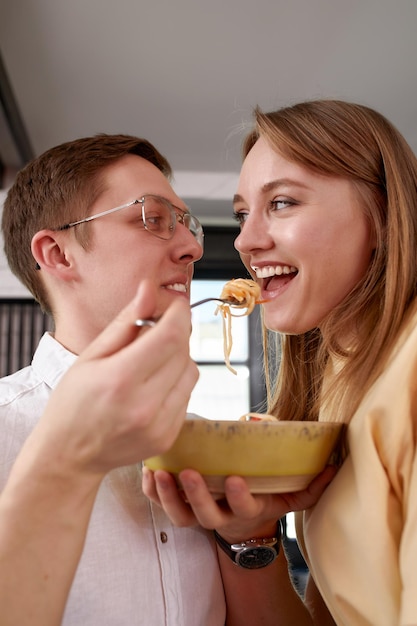 Lächelnder, liebevoller Ehemann, der Frau füttert, junges Paar, das einen zärtlichen Moment genießt, romantisches Date, das Spaß daran hat, japanische Küche zu essen, glückliche Familie, die ein freies Wochenende in der modernen Küche verbringt