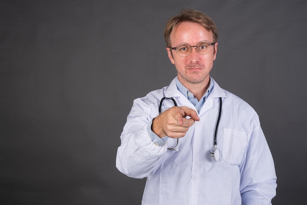Lächelnder männlicher Arzt mit Stethoskop im Arztkittel, der mit dem Finger auf grauen Hintergrund zeigt
