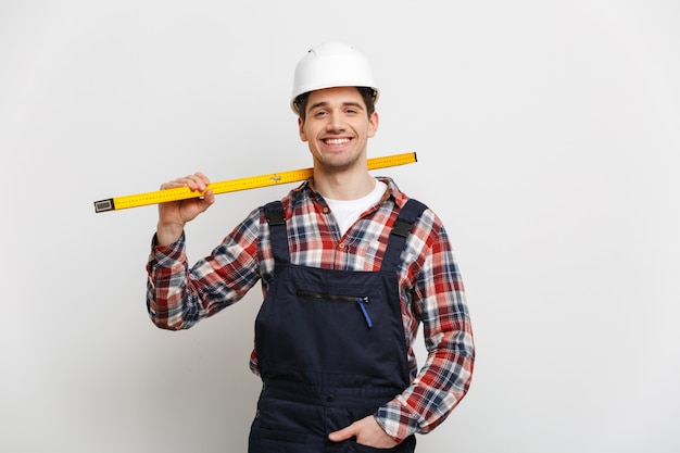 Lächelnder männlicher Baumeister im Schutzhelm, der ebenes Werkzeug über grauer Wand hält