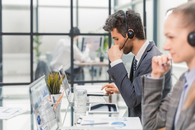 Lächelnder männlicher Callcenter-Betreiber mit Kopfhörern sitzt im modernen Büro mit Kollegen im Hintergrund und berät online