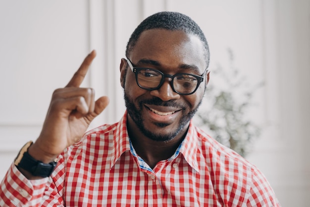 Lächelnder männlicher Englischlehrer der afrikanischen Ethnie in Gläsern hebt die Hand mit dem Zeigefinger nach oben