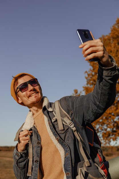 Lächelnder männlicher Rucksacktourist mit Sonnenbrille, der im Herbstwald ein Selbstporträt mit dem Smartphone macht