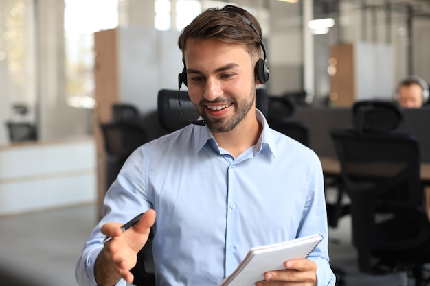 Lächelnder männlicher Unternehmensberater mit Kopfhörern im modernen Büro, Videoanruf mit Blick auf den Laptop-Bildschirm. Man Kundenservice-Support-Agent-Helpline im Online-Chat.