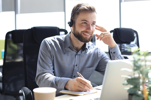 Lächelnder männlicher Unternehmensberater mit Kopfhörern im modernen Büro, Videoanruf mit Blick auf den Laptop-Bildschirm. Man Kundenservice-Support-Agent-Helpline im Online-Chat.