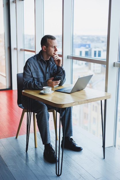 Lächelnder männlicher Unternehmer in Freizeitkleidung, der mit Laptop am Tisch sitzt und tagsüber auf dem Smartphone in einem modernen Arbeitsbereich mit großem Fenster spricht