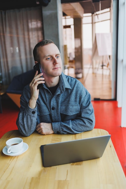 Lächelnder männlicher Unternehmer in Freizeitkleidung, der mit Laptop am Tisch sitzt und tagsüber auf dem Smartphone in einem modernen Arbeitsbereich mit großem Fenster spricht