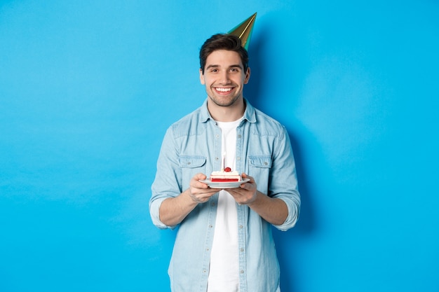 Lächelnder Mann, der b-Tageskuchen hält und Geburtstagsfeierhut trägt, der über der blauen Wand feiert