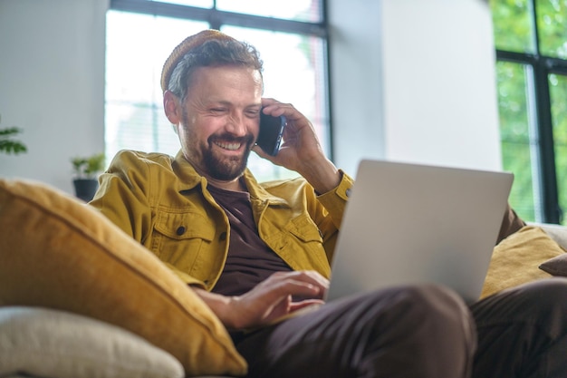 Foto lächelnder mann, der ein telefongespräch führt, während er am laptop auf dem sofa im büro arbeitet. er trug freizeitkleidung und war höchstwahrscheinlich programmierer oder it-experte