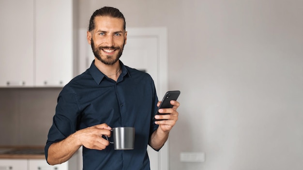 Lächelnder Mann, der eine Tasse Kaffee und ein Handy hält, während er in die Kamera schaut