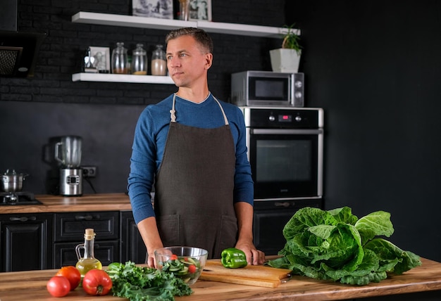 Lächelnder Mann, der Salat kocht, frisches Gemüse in moderner schwarzer Küche schneidet