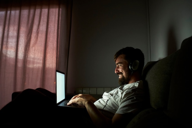 Lächelnder Mann, der spät zu Hause arbeitet. Er sitzt im Dunkeln auf der Couch.