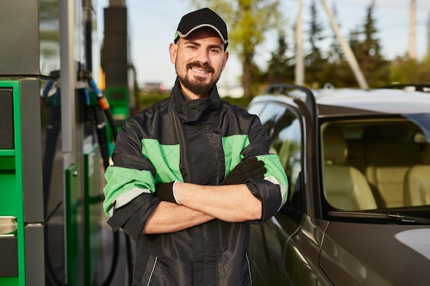 Lächelnder Mann, der während der Arbeit an einer Tankstelle in der Nähe eines modernen Autos steht