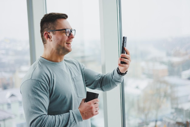 Lächelnder Mann in Freizeitkleidung, der mit Kaffee am Fenster steht und tagsüber in einem modernen Arbeitsplatz mit großem Fenster auf das Smartphone schaut