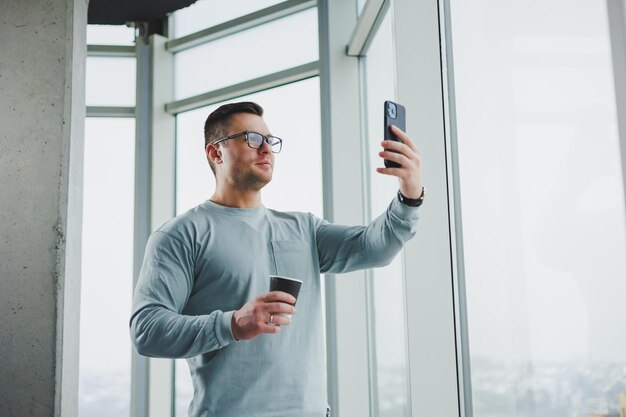 Lächelnder Mann in Freizeitkleidung, der mit Kaffee am Fenster steht und tagsüber in einem modernen Arbeitsplatz mit großem Fenster auf das Smartphone schaut