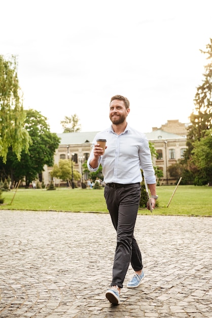 Lächelnder Mann in voller Länge in formeller Kleidung, der durch Park geht und Kaffee zum Mitnehmen und silbernen Laptop trägt