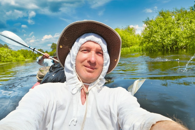 Foto lächelnder mann in weißem hoodie und hut, der während der sommerlichen flussabenteuerreise ein selfie-foto auf dem kajak macht