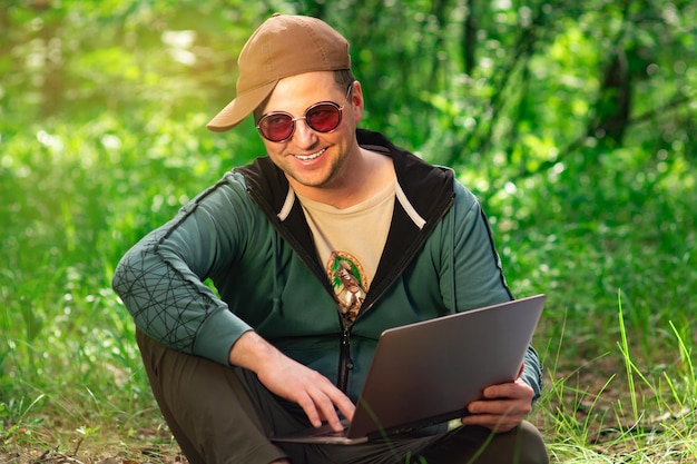 Lächelnder Mann mit roter Sonnenbrille und Baseballkappe, der auf dem Gras im Wald sitzt und einen Laptop hält