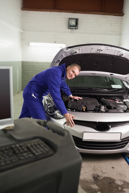 Lächelnder Mechaniker, der auf einem Auto nahe bei einem Computer sich lehnt