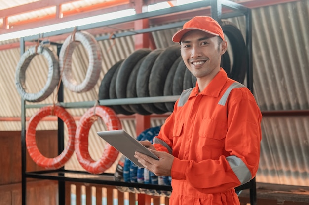 Lächelnder Mechaniker im Wearpack mit einem digitalen Tablet in der Werkstatt mit einem Reifenständer dahinter