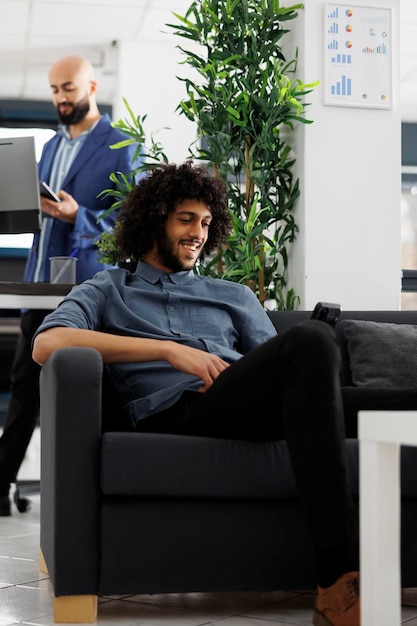 Lächelnder Mitarbeiter eines Start-up-Unternehmens schaut sich während einer Arbeitspause im Geschäftsbüro ein Video auf dem Smartphone an. Ein fröhlicher arabischer Unternehmer benutzt sein Handy und entspannt sich im Coworking-Bereich