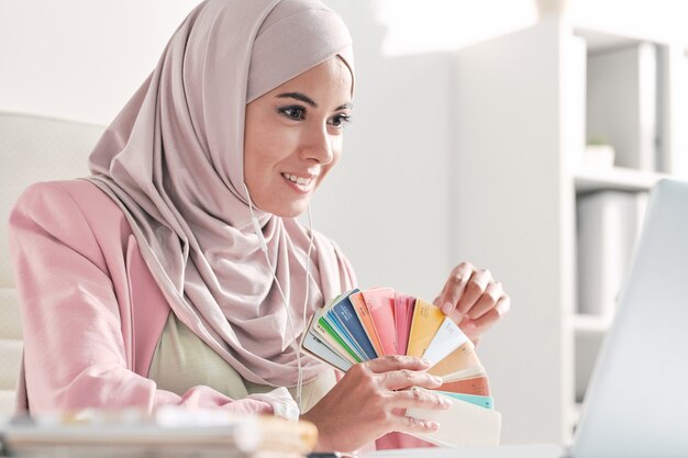 Lächelnder muslimischer Designer im rosa Hijab, der dem Kunden Farbmuster zeigt, während er über Videokonferenz-App mit ihm spricht