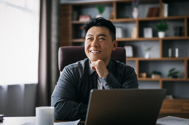 Lächelnder nachdenklicher erwachsener asiatischer Geschäftsmann, inspiriert von der Idee am Laptop zum Arbeitsplatz im Innenbereich des Home Office