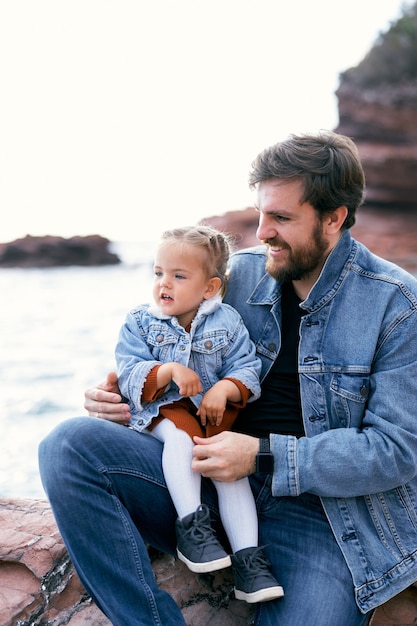 Lächelnder Papa mit einem kleinen Mädchen im Arm sitzt an einem felsigen Ufer vor der Kulisse der Berge