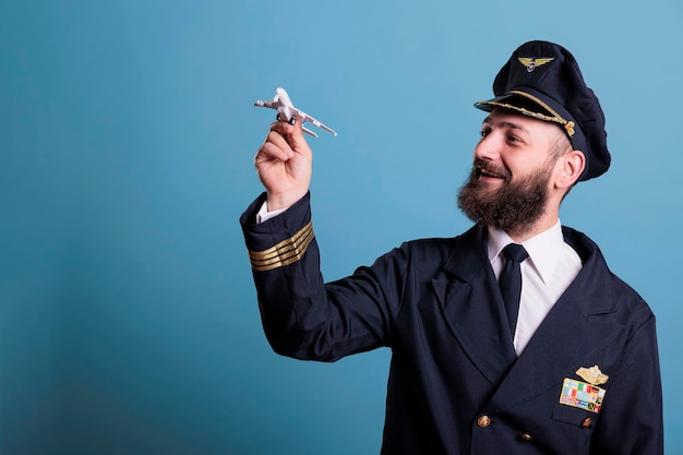 Lächelnder Pilot in Uniform mit Flugzeugmodell, Flieger der Luftfahrtakademie, der mit dem Flugzeug spielt. Flugzeugbesatzungsmitglied, das kommerzielles Jet-Spielzeug betrachtet, Seitenansicht, Studio, mittlerer Schuss