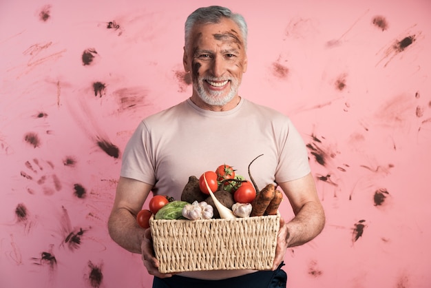 Lächelnder, positiver älterer Mann, der einen Korb des frischen Gemüses gegen eine schmutzige rosa Wand hält