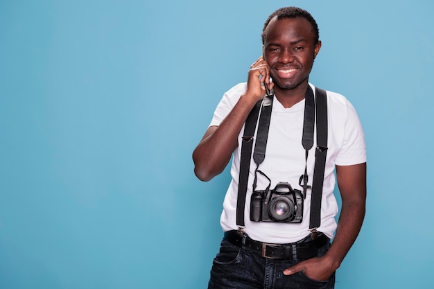 Lächelnder professioneller Fotograf, der auf dem Smartphone spricht, während er auf blauem Hintergrund steht. Selbstbewusster junger Fotografie-Enthusiast mit DSLR-Kamera beim Sprechen auf einem modernen Touchscreen-Telefongerät.