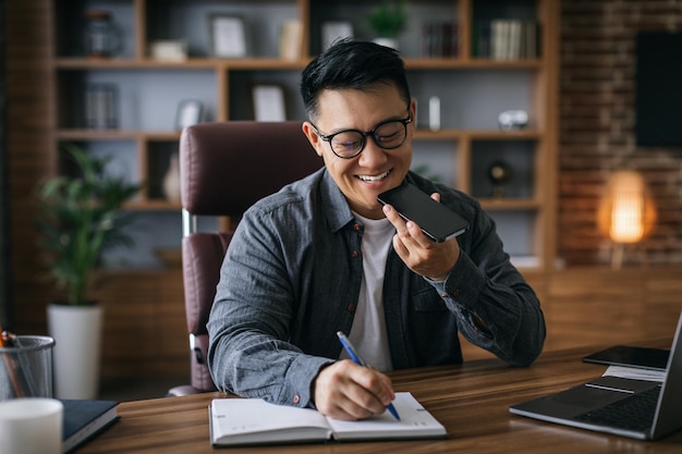 Lächelnder reifer asiatischer Geschäftsmann mit Brille, der auf dem Smartphone spricht und sich am Arbeitsplatz zu Hause Notizen macht