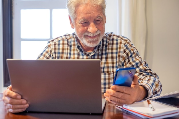 Lächelnder reifer bärtiger Mann, der mit Laptop und Büchern am Tisch sitzt und dem Online-Kurs folgt Älterer Mann in kariertem Hemd, der Unterrichtstätigkeit genießt und mit neuen Technologien lernt
