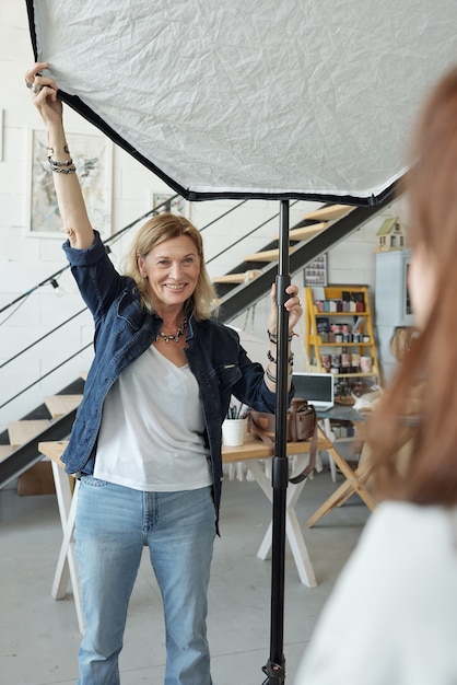 Lächelnder reifer Fotograf in der Jeansjacke, die Softbox vor dem Fotoshooting im Studio anpasst