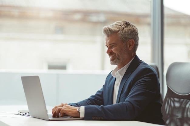 Lächelnder reifer Geschäftsmann, der am Laptop arbeitet, während er im modernen Coworking sitzt
