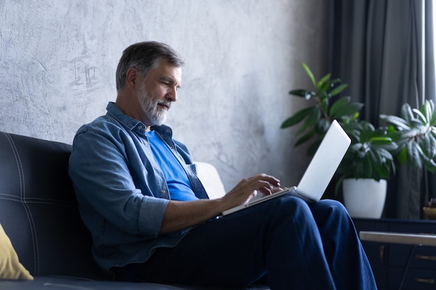 Lächelnder reifer Mann sitzt auf einem Sofa und tippt auf einem Laptop in einem Wohnzimmer. Arbeiten von zu Hause aus im Quarantäne-Lockdown. Soziale Distanzierung Selbstisolation