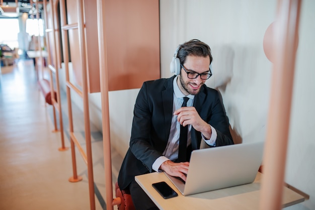 Lächelnder schöner kaukasischer bärtiger Geschäftsmann im Anzug, mit Brille und Kopfhörern, die am Arbeitsplatz sitzen und Laptop verwenden.