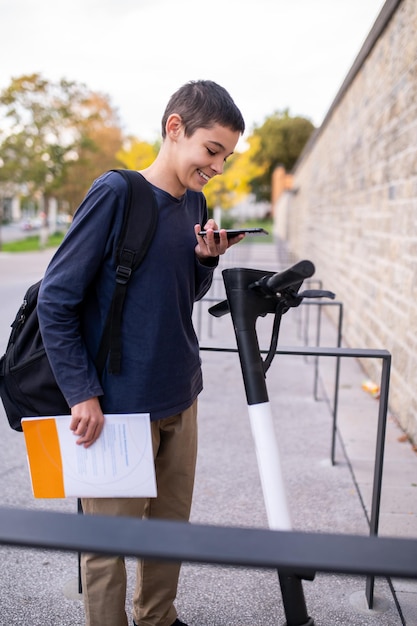 Lächelnder Schuljunge, der auf dem E-Scooter-Deck steht und sein Handy über den LCD-Bildschirm hält