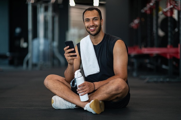 Lächelnder schwarzer männlicher Athlet, der sich nach dem Training im Fitnessstudio mit dem Smartphone entspannt