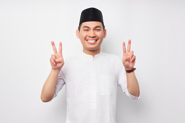 Lächelnder selbstbewusster junger asiatischer muslimischer Mann, der in die Kamera schaut und das Siegeszeichen zeigt, das auf weißem Hintergrund isoliert ist Menschen religiöses islamisches Lebensstilkonzept
