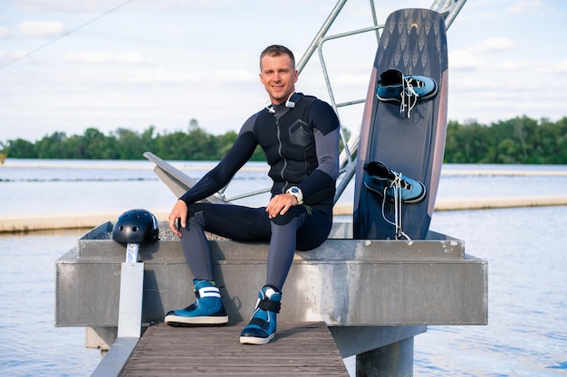 Lächelnder selbstbewusster Wakeboarder, der sich nach dem Training auf dem Pier mit Wakeboard ausruht