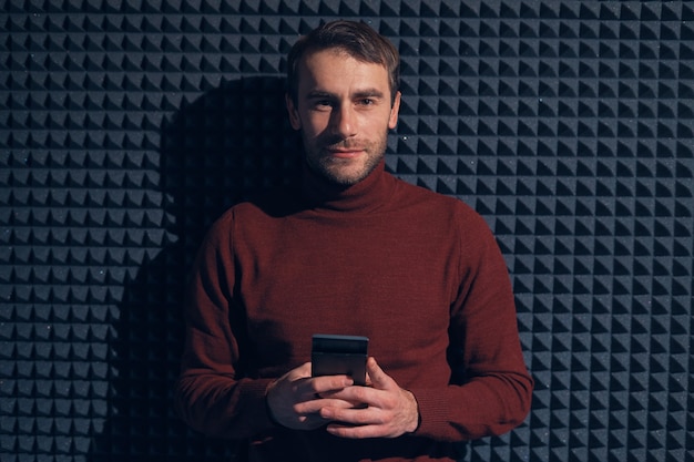 Lächelnder sicherer Mann, der Smartphone über grauem Hintergrund mit geometrischen Formen und Lichtspiel hält.