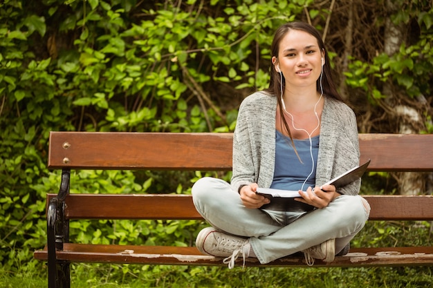 Lächelnder Student, der auf hörender Musik der Bank mit Handy sitzt und Buch im Park an schoo hält