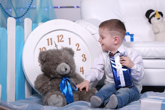 Lächelnder süßer Junge, der sein Geschenk während der Weihnachtszeit hält. große weiße Uhr und Teddybär an der Wand