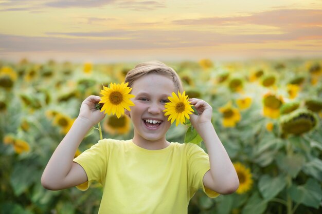 Lächelnder süßer Junge, Teenager, der auf einem Feld steht und zwei Sonnenblumen in der Nähe hält. Sommerferien, glückliche Kindheit