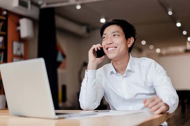 Lächelnder tausendjähriger asiatischer Geschäftsmann, der am Telefon im Firmenbüro mit Laptop spricht