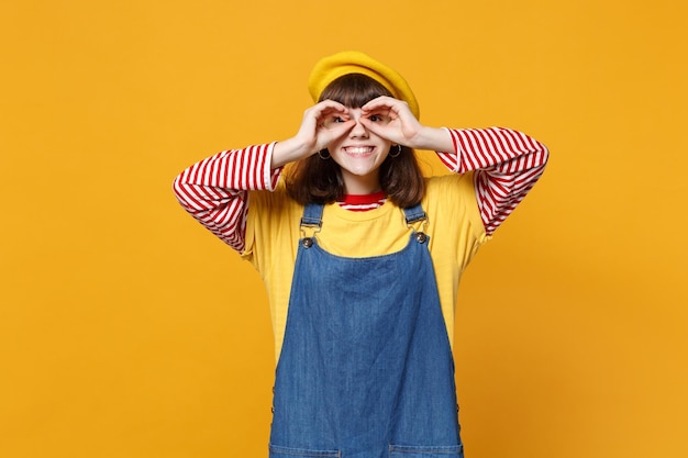 Lächelnder Teenager in französischer Baskenmütze und Denim-Sommerkleid, der die Hände in der Nähe der Augen hält und eine Brille oder ein Fernglas isoliert auf gelbem Hintergrund imitiert. Menschen Emotionen, Lifestyle-Konzept. Mock-up-Kopienbereich.