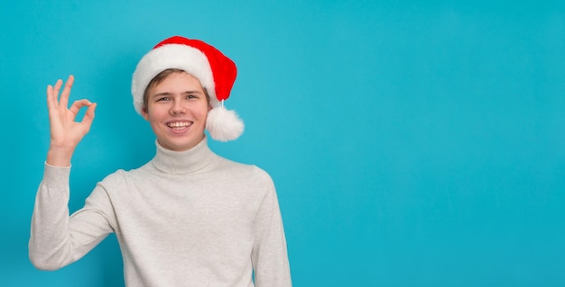 Lächelnder Teenager-Junge mit Weihnachtsmütze, der auf blauem Hintergrund eine OK-Geste zeigt