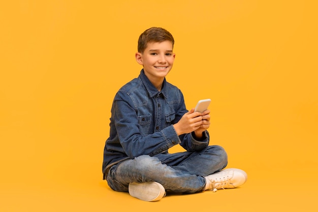 Lächelnder Teenager mit Smartphone in den Händen sitzt mit gekreuzten Beinen auf gelbem Hintergrund