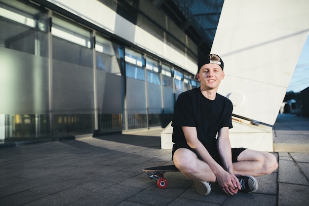 lächelnder Teenager-Skater sitzt auf einer Tafel auf dem Hintergrund der modernen Architektur. Skater ruht auf einem Brett zum Skifahren.