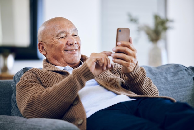 Lächelnder Tipper und ein älterer Mann mit einem Telefon auf dem Sofa für Social-Media-Kontakte oder Kommunikation. Fröhliche Entspannung und ein älterer Mensch auf der heimischen Couch mit einem Handy für eine App oder zum Lesen eines Chats
